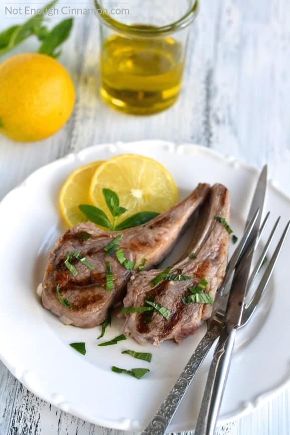 overhead shot of lemon and mint marinated pan-fried lamb chops on a white plate with cutlery on the side