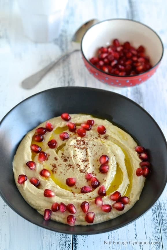 smooth homemade hummus served in a black bowl with pomegranate seeds and sumac sprinkled on top and a small dish with more pomegranate arils in the background