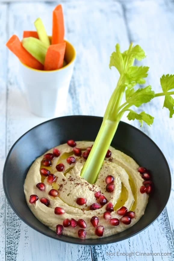 creamy and smooth homemade hummus served in a black bowl sprinkled with sumac and pomegranate arils, with some veggie sticks on the side - easy hummus recipe 