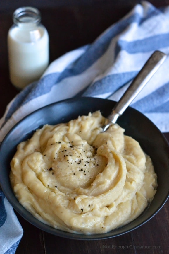 Easy Slow Cooker Mashed Potatoes with goat cheese served in a black bowl with some cracked pepper on top and small milk bottle in the back