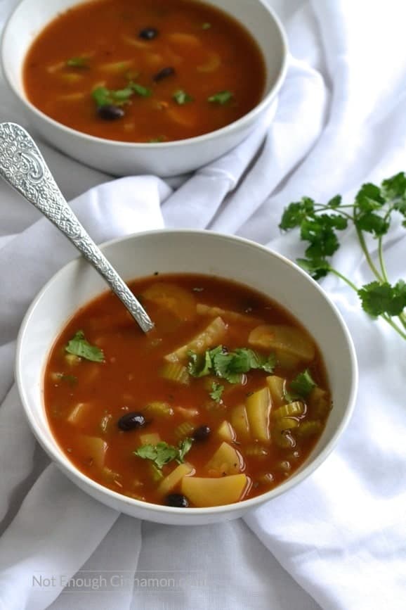 two bowls of veggie soup with chunks of zucchini, celery and black beans in a healthy tomato broth with a twig of parsley on the side