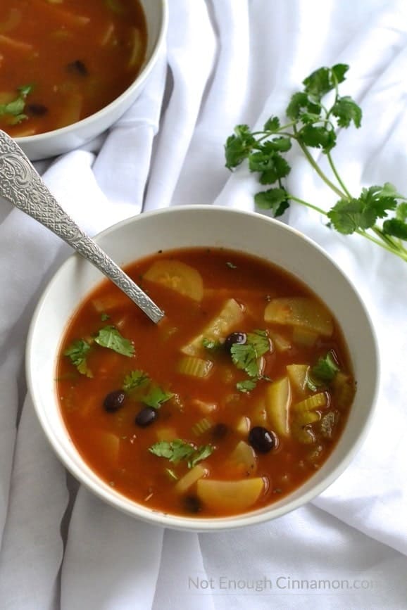 overhead shot of a white bowl with a tomato-based veggie soup with sliced zucchini, black beans and slivers on celery 