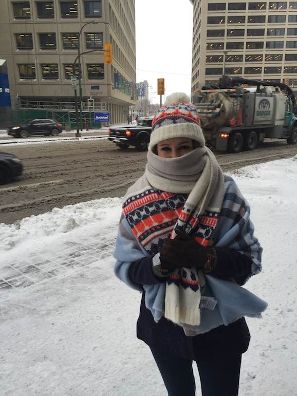 a woman standing on a snow-covered sidewalk outside of Winnipeg station wrapped in several shawls