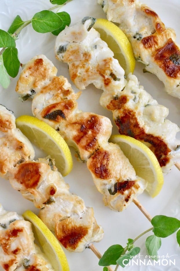 close-up of Greek Lemon Chicken Skewers arranged on a white serving plate with fresh oregano and lemon slices