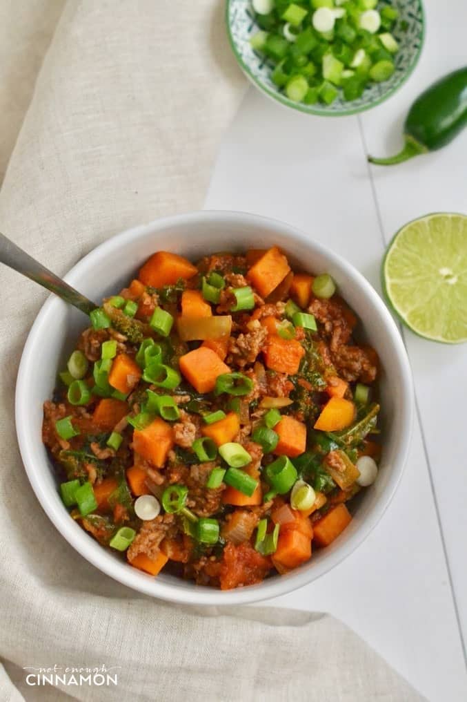 overhead shot of paleo no bean sweet potato chili with beef and kale in a white bowl with jalapenos, scallions and lime on the side