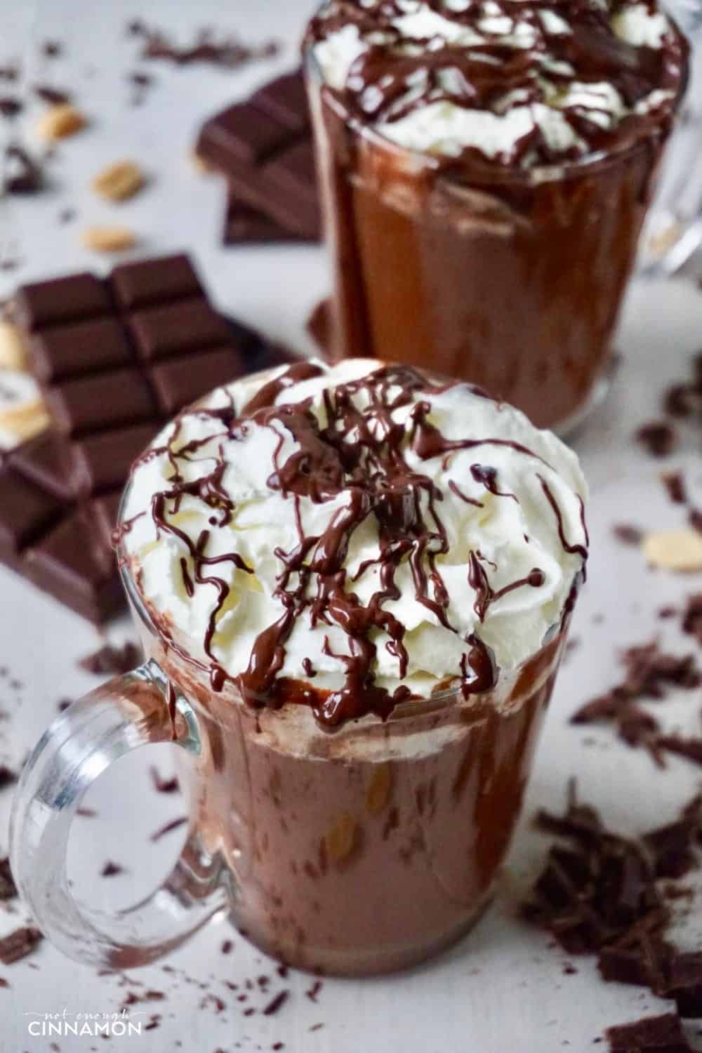 two mugs of healthier peanut butter hot chocolate topped with whipped cream and chocolate sauce