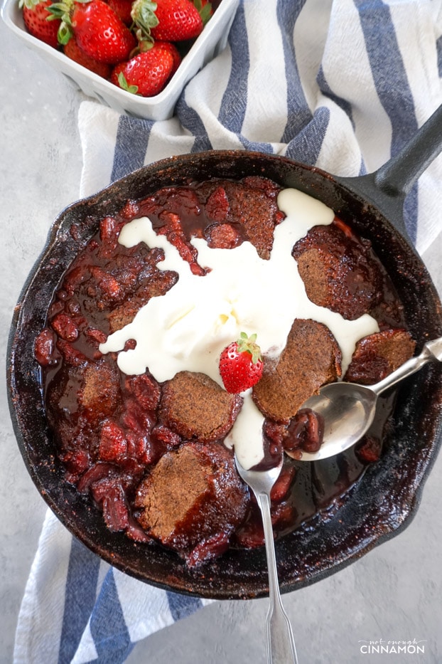 Strawberry Cobbler in a cast iron skillet, with two spoons and melting ice cream scoops on top.