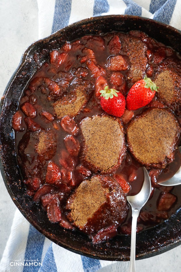 Strawberry cobbler in a cast iron skillet over a white and blue stripped dish towel, with two spoons and two fresh strawberries on top. 