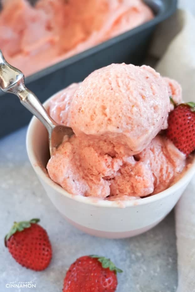Several scoops of strawberry banana ice cream in a white and pink small bowl, being spooned with a metal spoon.