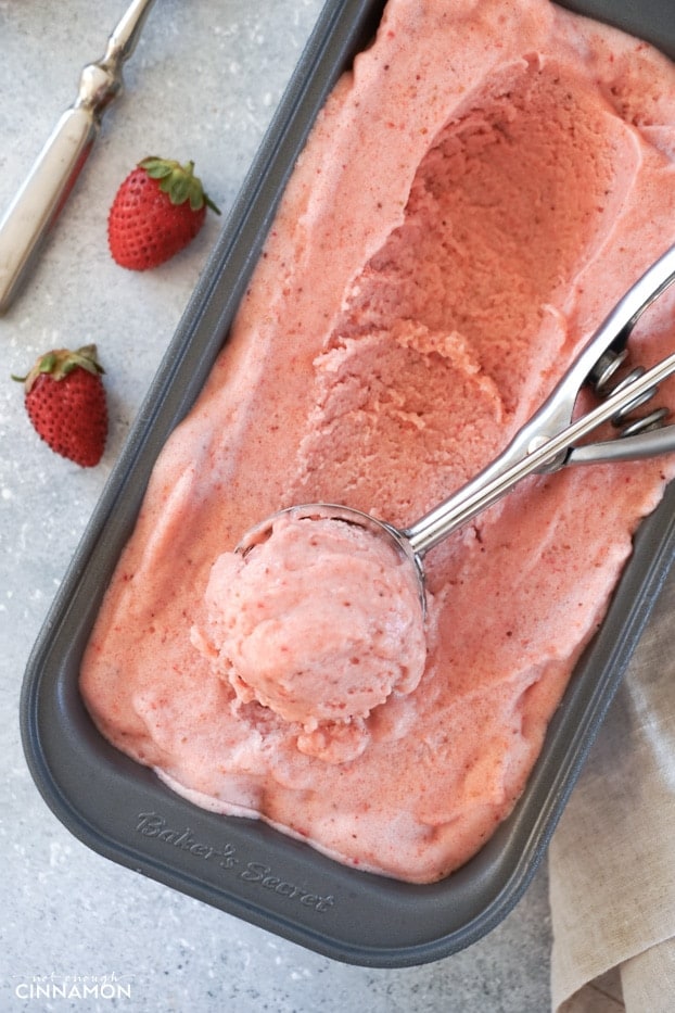 Strawberry banana ice cream in a metal tin being scooped with an ice cream scoop, with a spoon and fresh strawberries in the background. 