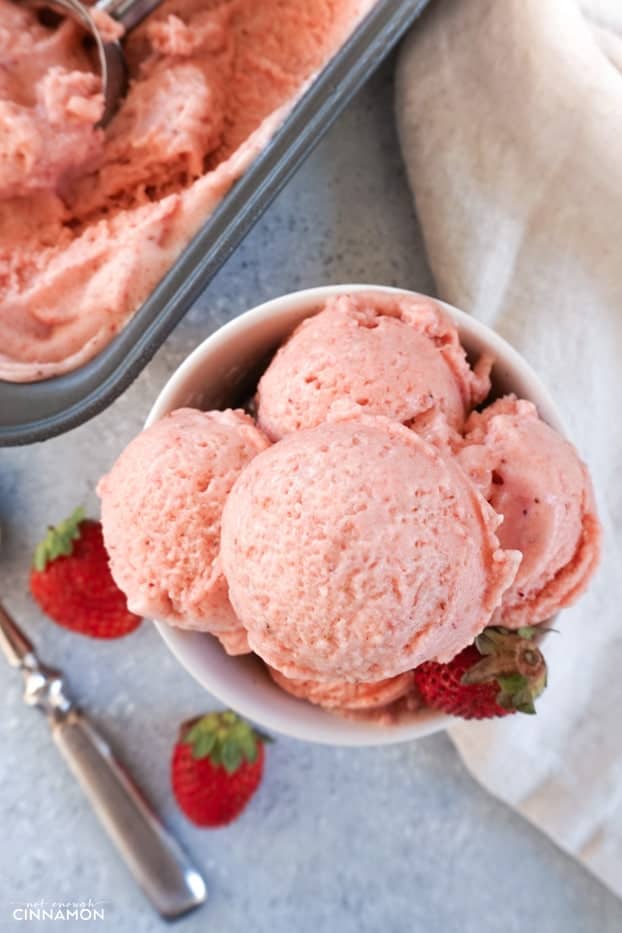 Strawberry banana ice cream in a bowl, with a spoon, fresh strawberries, a napkin and the ice cream container is the background.
