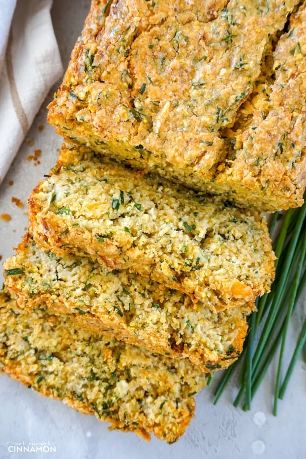 A loaf of cheddar chive zucchini bread, cut partially in three slices, with a few chive sprigs on the side and a striped brown and white napkin