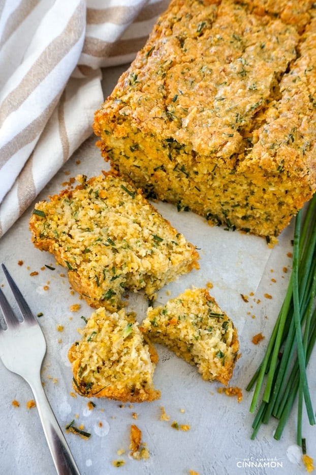 A slice of cheddar chive zucchini bread broken in pieces, next to a fork, with the bread loaf in the background and fresh chives on the side.