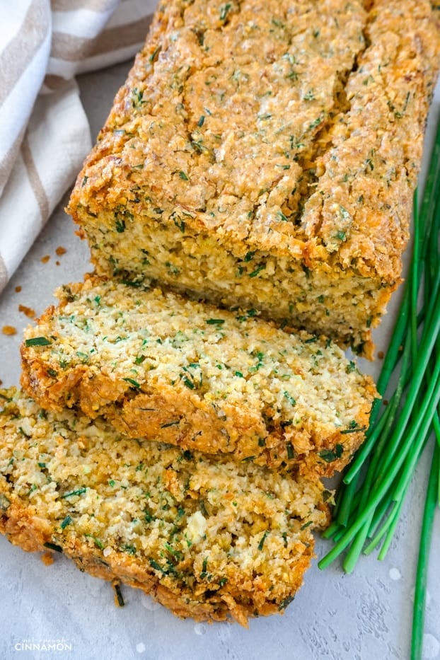 A loaf of cheddar chive zucchini bread, cut partially in two slices, with a few chive sprigs on the side and a striped brown and white napkin 