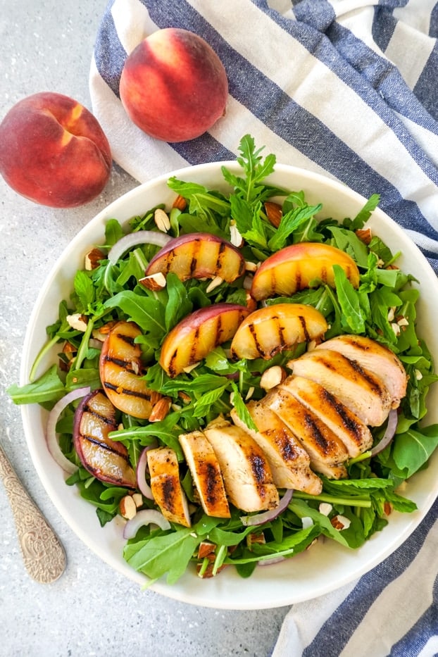 Grilled chicken slices and grilled peach slices on a bed of arugula and fresh herbs i a large shallow bowl, with a blue and white striped towel and two peaches in the background.
