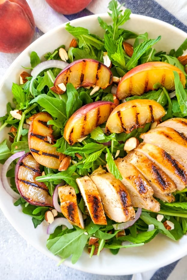 Sliced grilled chicken and grilled peaches, on a bed of arugula and fresh herbs, with red onion slices and chopped almonds in a large white shallow plate.
