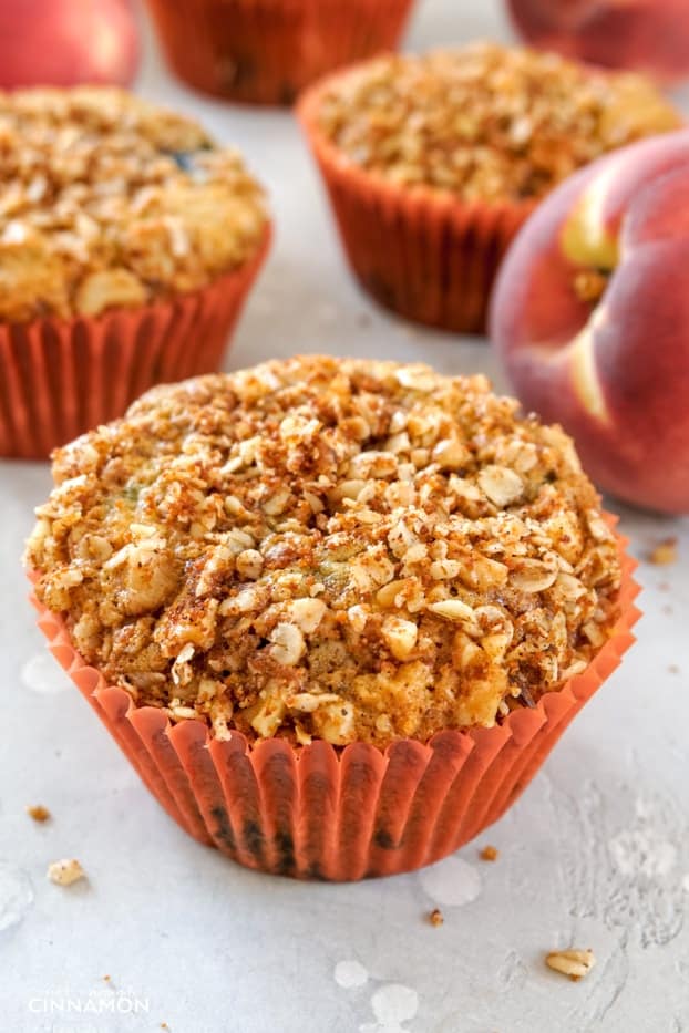 A peach and blueberry muffin with a streusel topping in an orange liner, with a peach and other muffins in the background.