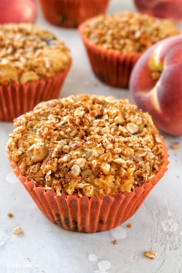 A peach and blueberry muffin with a streusel topping in an orange liner, with a peach and other muffins in the background. 