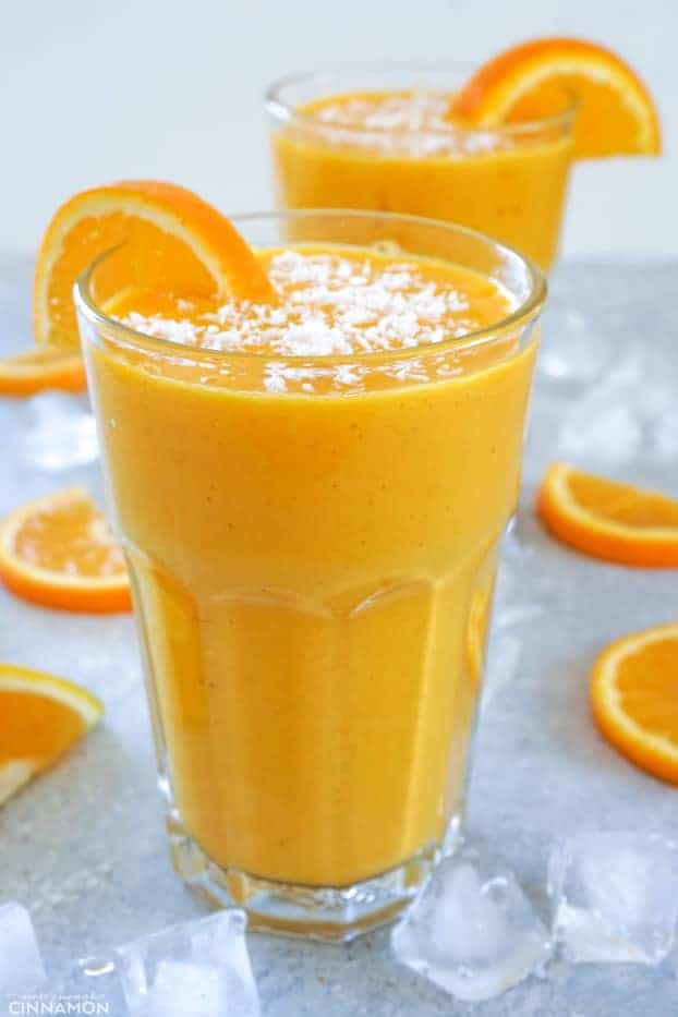 A tall glass of smoothie surrounded by orange slices and ice cubes