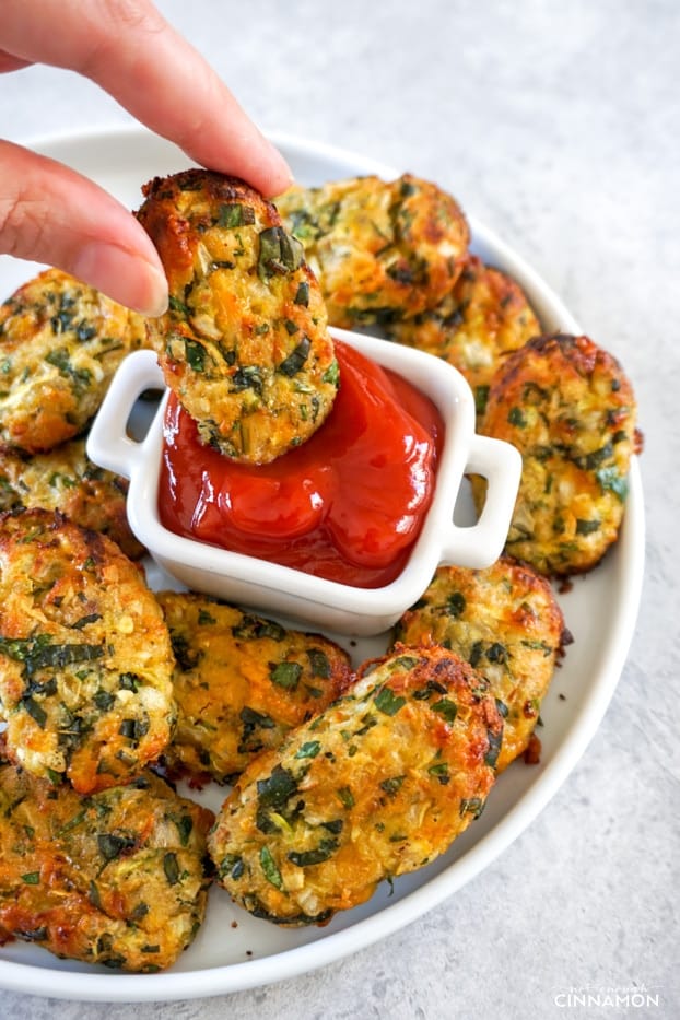 A plate of zucchini tater tots with a hand dipping a tot in ketchup.