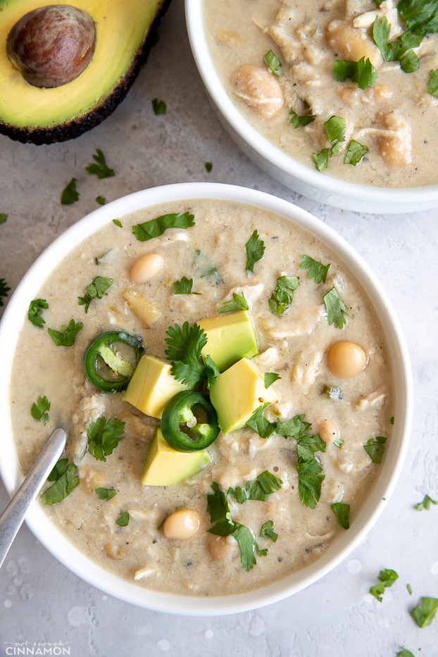 overhead shot of a bowl of slow cooker white chicken chili with a spoon, jalapeno slices and avocado cubes 