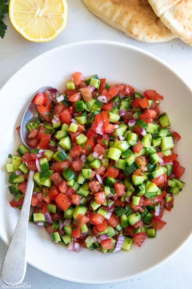 overhead shot of a bowl of Israeli salad with pita bread and lemon in the background 