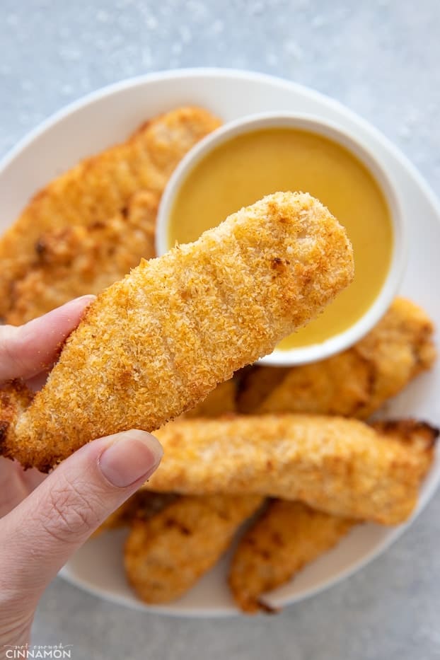 a baked coconut trusted chicken tender being held over a small bowl with whole30 honey mustard dip
