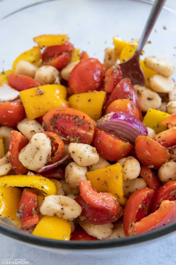 gnocchi, chopped bell pepper, onion, and tomatoes being tossed in a bowl with olive oil and herbs